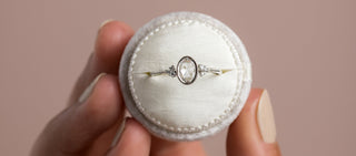Photo features a model holding a round ring box with a Rose Cut Melanie Casey ring inside, all in front of a pink background.