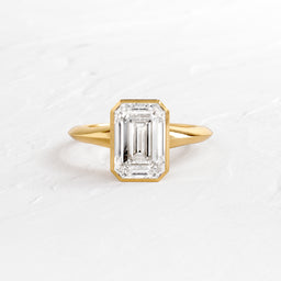 Embankment Ring in Emerald Cut Engagement Ring in Yellow Gold by Melanie Casey shown on a textured white background