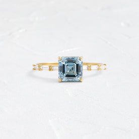 The Melanie Casey Fluency Ring in yellow gold with a 1.52ct. Lab-Grown Blue Diamond center stone shown on a textured white background.