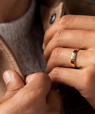 Photo showcases a model holding open the color of his brown jacket, showing a gray sweater underneath, with the Melanie Casey Hammered Mens Ban on one of his hands.