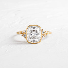 Snowdrift Bezel Set Ring Cushion Cut Engagement Ring in Yellow Gold by Melanie Casey shown on a textured white background