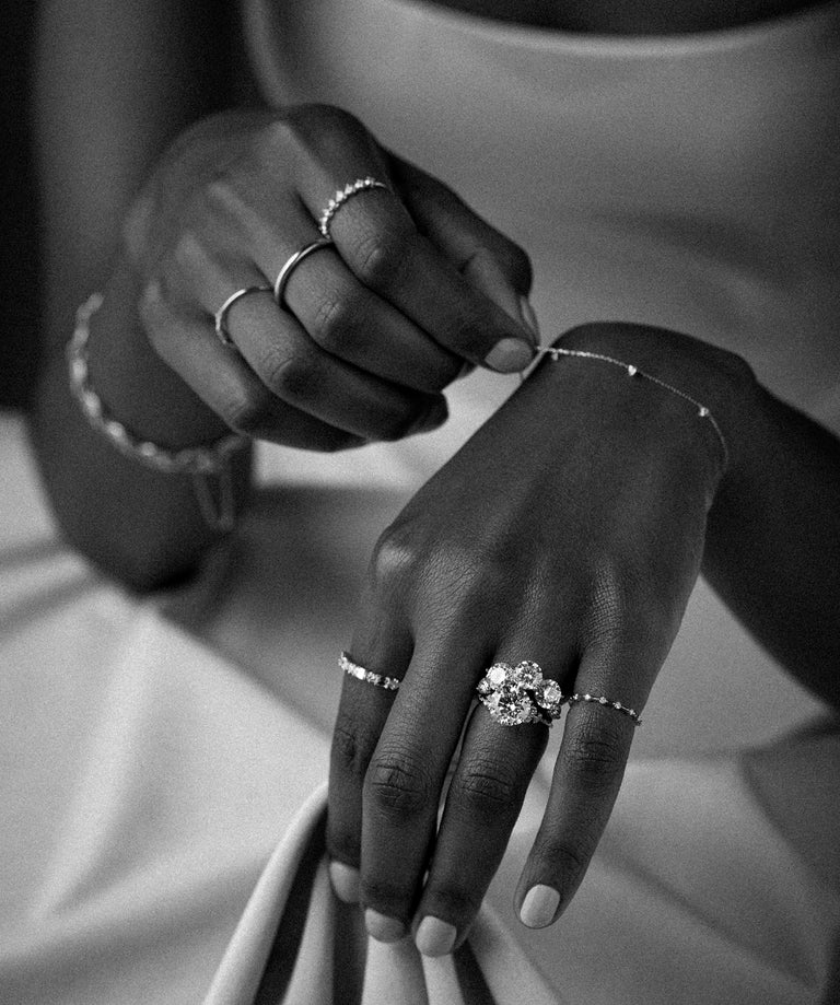 This black and white photo shows a model wearing an assortment of Melanie Casey jewelry including a round cut engagement ring, a mix of bands, and bracelets. She is in a bridal gown, touching the fabric of her gown with one hand and one of her bracelets with the other.