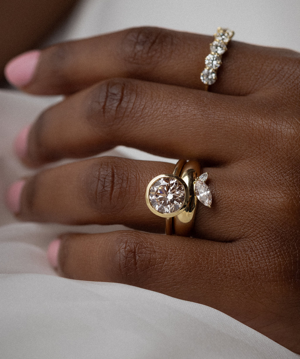 This photo features a model wearing a bezel-set round cut Melanie Casey engagement ring with a thick gold wedding band.