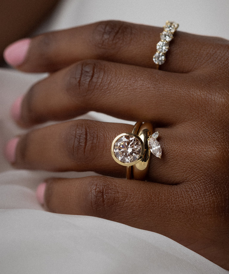 This photo features a model wearing a bezel-set round cut Melanie Casey engagement ring with a thick gold wedding band.