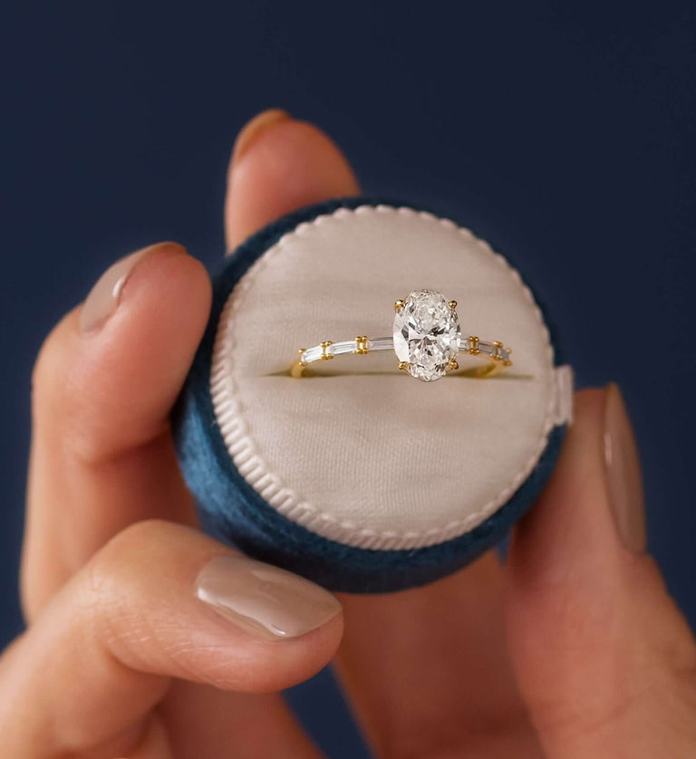 This photo features a model holding up a round velvet blue ring box with a Melanie Casey 14k yellow gold Fluency Engagement Ring in the center against a navy blue background.