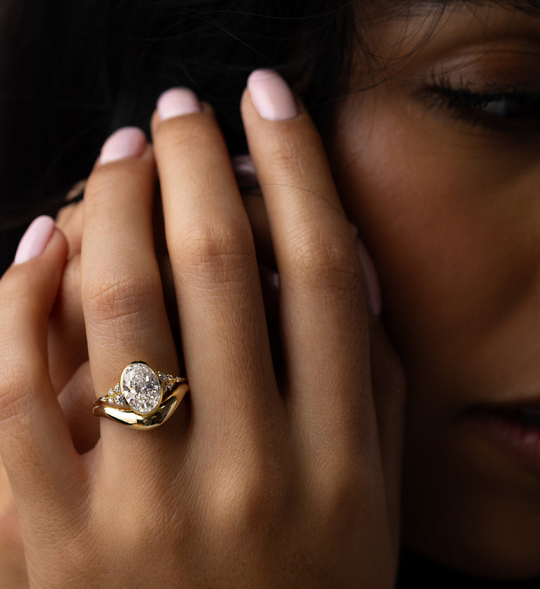 Photo showcases a model wearing the Oval Cut Bezel Set Snowdrift Engagement Ring on her hand, with her hand raised up by her face and her eyes looking downward.