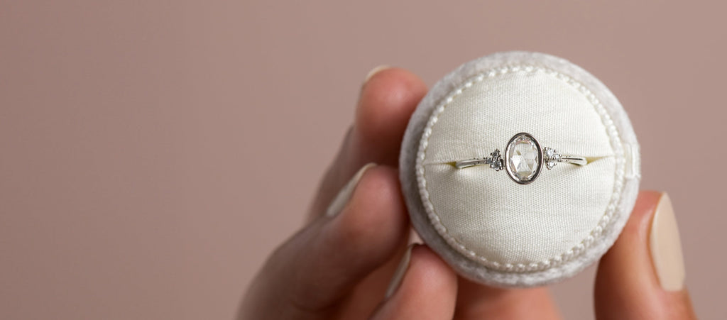 Photo features a model holding a round ring box with a Rose Cut Melanie Casey ring inside, all in front of a pink background.