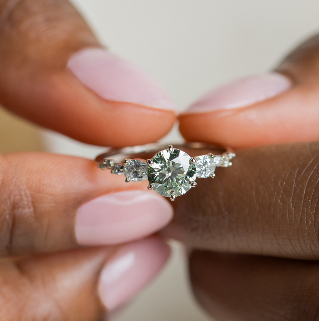 Photo showcases the Melanie Casey Wingspan Ring with a green diamond center stone being held by a model.