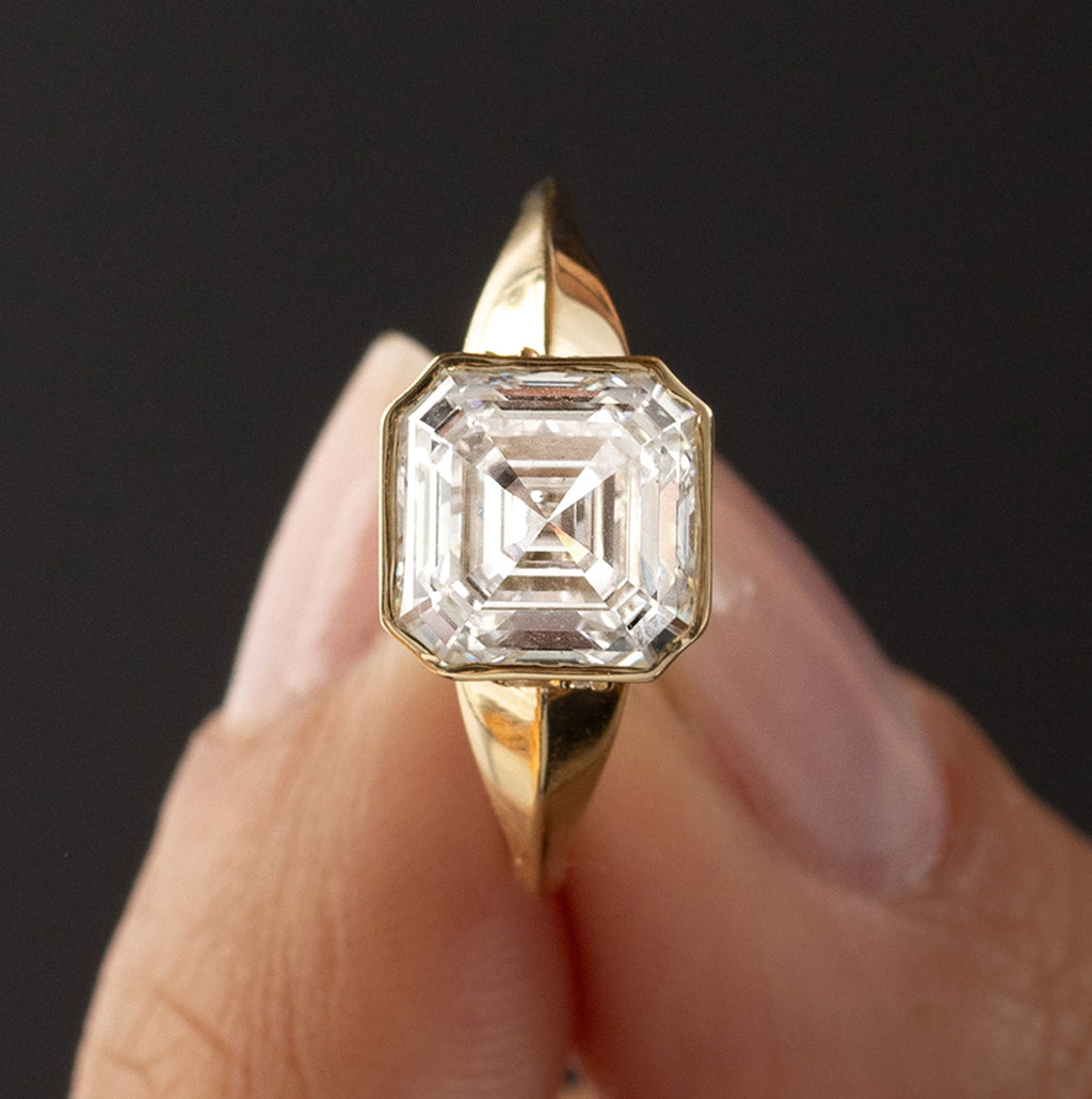 The Melanie Casey Bezel Set Elixir Ring in Asscher Cut being held up by a model against a dark background.