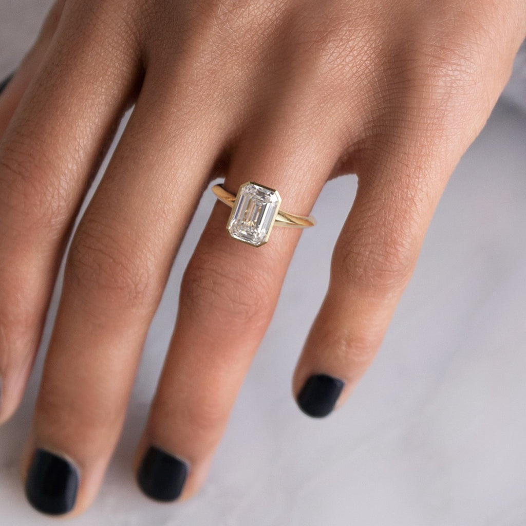 Photo features a model wearing the Melanie Casey Bezel Set Embankment Ring in Emerald Cut.