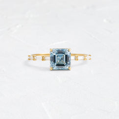 The Melanie Casey Fluency Ring in yellow gold with a 1.52ct. Lab-Grown Blue Diamond center stone shown on a textured white background.