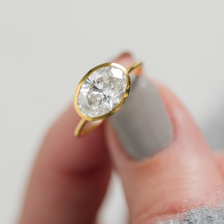 Photo features a model holding the Melanie Casey Bezel Engagement Oval Cut Formation Ring in Yellow Gold.