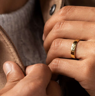 Photo showcases a model holding open the color of his brown jacket, showing a gray sweater underneath, with the Melanie Casey Hammered Mens Ban on one of his hands.