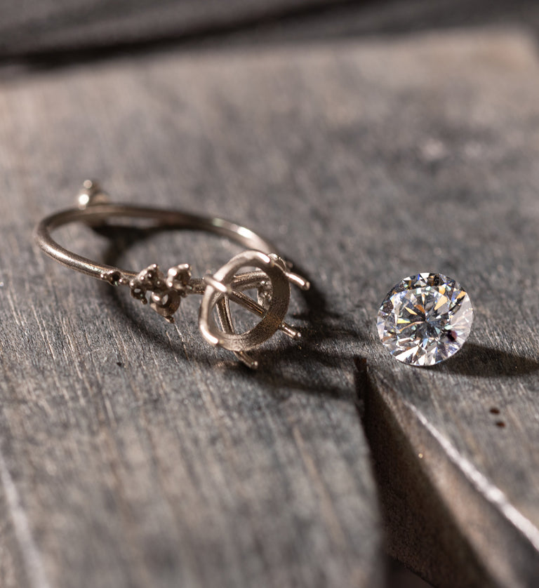 A photo of a round diamond sitting next to the Melanie Casey ring setting it will be set into on the bench.