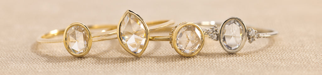 Four bezel set rose cut Melanie Casey rings, sitting on top of a pinkish tan textured surface in a straight line, one slightly on top of the other.