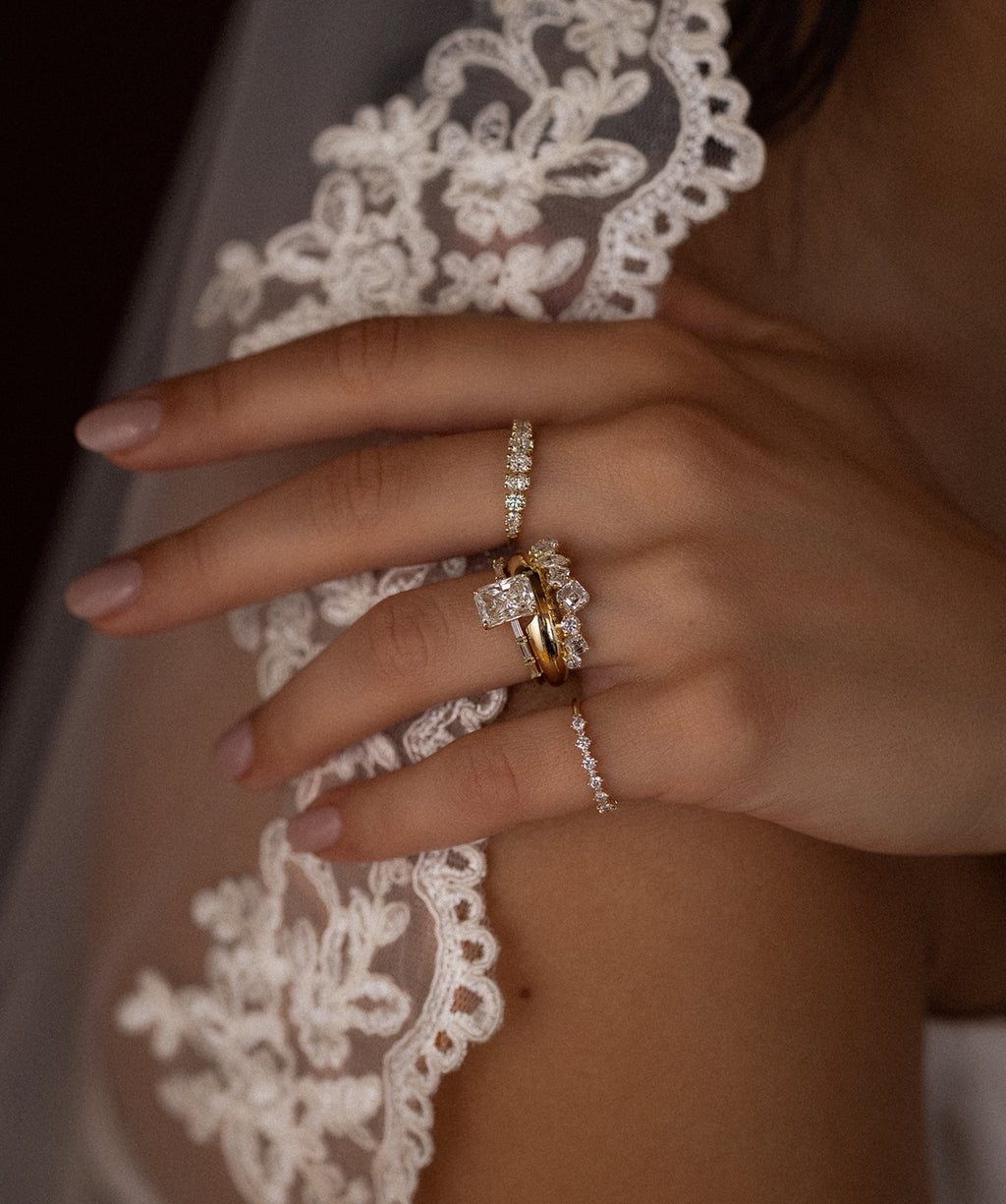 This photo showcases a model wearing a stack of Melanie Casey rings. She is wearing a lacy wedding veil, and her hand is placed on her shoulder, showing off the rings.