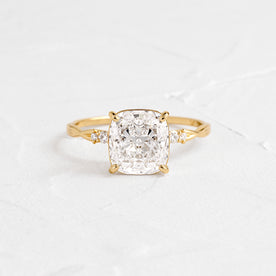 Trestle Cushion Cut Ring Engagement Ring in Yellow Gold by Melanie Casey shown on a textured white background