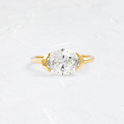 Stanza Ring in Oval Cut Engagement Ring in Yellow Gold by Melanie Casey shown on a textured white background